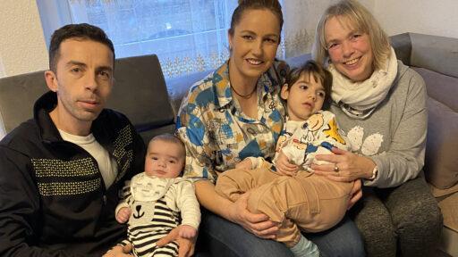 Eine junge Familie sitzt mit ihren Babies auf dem Schoß auf einem Sofa im Wohnzimmer mit einer Löwenherz-Ehrenamtlichen