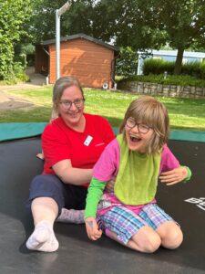 Eine Pflegekraft von Löwenherz und ein lachendes Mädchen sitzen auf einem Trampolin