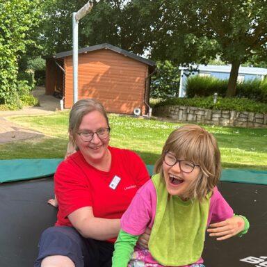Eine Pflegekraft von Löwenherz und ein lachendes Mädchen sitzen auf einem Trampolin