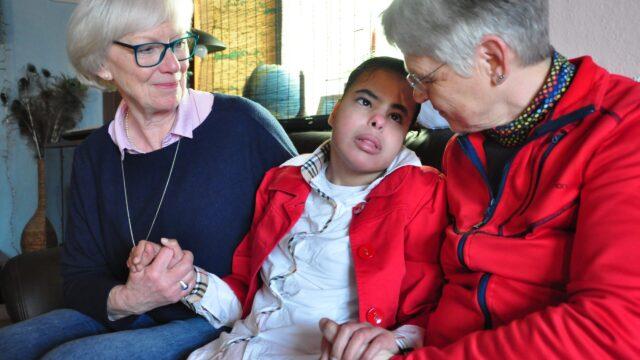 Zwei Löwenherz-Ehrenamtliche sitzen mit einer pflegebedürftigen jungen Frau auf einem Sofa