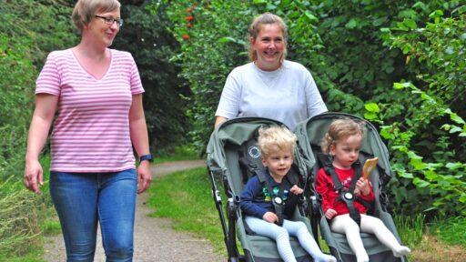 Eine junge Frau schiebt einen Doppel-Kinderwagen mit zwei Kindern, die Mutter geht daneben und lacht