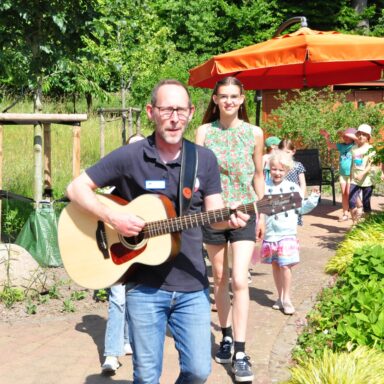 Ein Mann mit Gitarre marschiert vor einem Kinderchor durch den Löwenherz-Garten.
