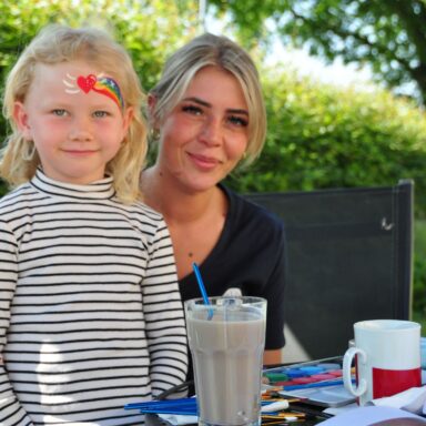 Ein kleines, blondes Mädchen mit geschminktem Gesicht und der schminkenden Frau im Hintergrund