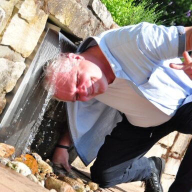 Ein Mann kniet am Boden und hält seinen Kopf unter einen kleinen Wasserfall im Löwenherz-Garten.