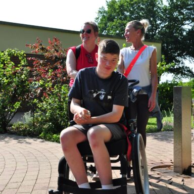 Zwei Frauen und ein Jugendlicher im Rollstuhl neben einer Wasserstehle.