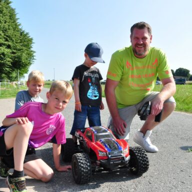 Ein Mann und drei Kinder posieren neben einem großen, ferngesteuerten Auto auf einer Straße.