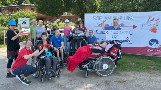 Eine Gruppe von Pflegekräften, Eltern und erkrankten Kindern steht vor einem Bauzaun vor dem Kinderhospiz Löwenherz