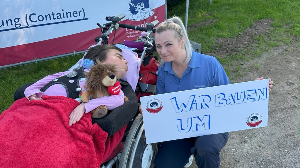 Eine Pflegekraft kniet mit einem Schild neben einer Jugendlichen im Rollstuhl vor einem Bauzaun.