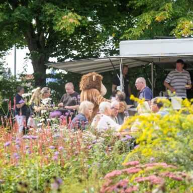 Menschen an einer Bratwurstbude, im Vordergrund der blühende Löwenherz-Garten