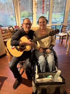 Musik-Therapeut Sören Tesch sitzt mit Gitarre auf dem Knie neben Johanna, einer jungen Frau im Rollstuhl.