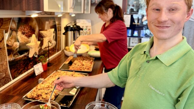 Ein junger Mann gibt steht an einem Büfett im Kinderhospiz Löwenherz und gibt Salat in eine Schüssel. Im Hintergrund bereitet seine Kollegin Pizza auf einem Blech vor.
