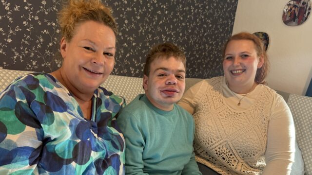 Ein junger Mann sitze auf einem Sofa, neben ihm seine Mutter und seine Tochter.