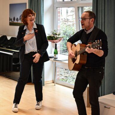 Sören Tesch musiziert im Stehen mit einer Gitarre, daneben steht Gaby Letzing und lacht.