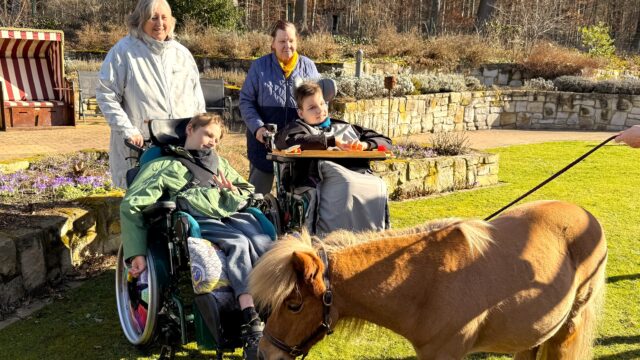 Zwei Großmütter stehen hinter ihren Enkeln, die im Rollstuhl sitzen, im Garten von Löwenherz. Im Vordergrund steht ein Pony.